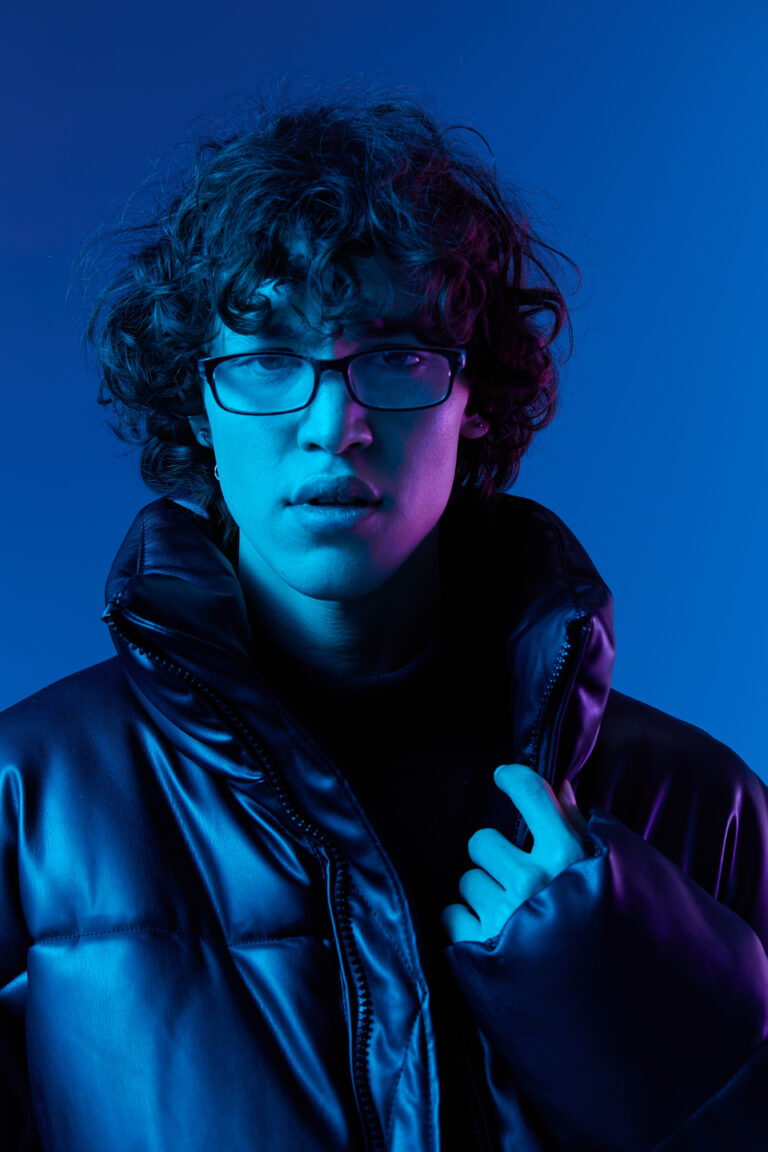 Man in winter jacket model poses in glasses smile, hipster lifestyle, portrait blue background, mixed neon light, fashion style and trends boys teenager, copy space. High quality photo