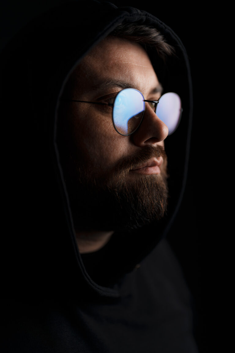 Side profile of a man with a hood, his glasses catching a glimpse of light in the dark