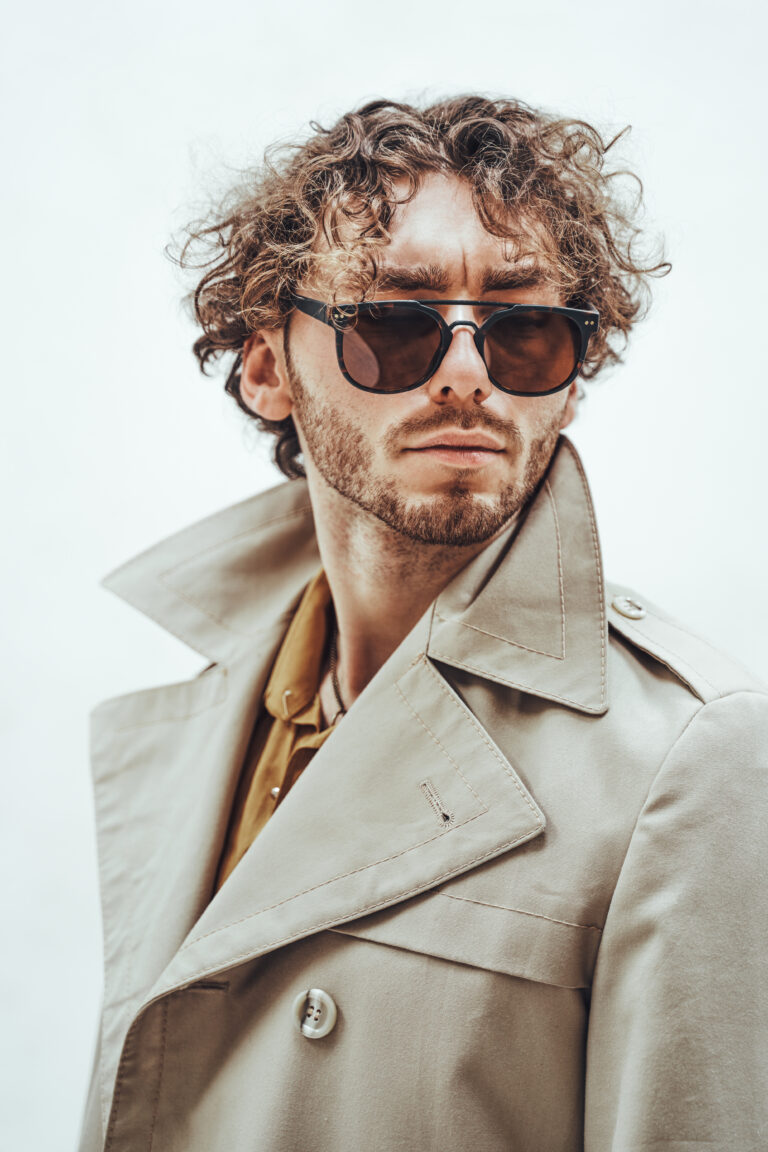 Stylish and rich young man posing for the photoshoot on the white background, looking assertive while wearing sunglasses and trench coat